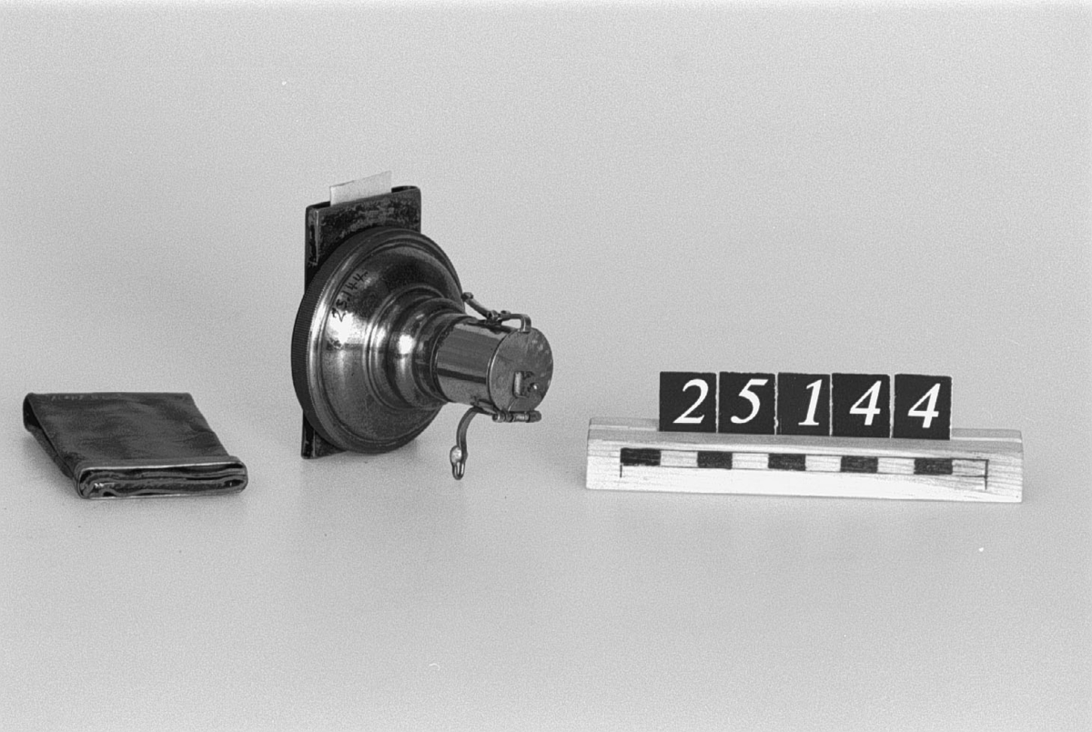 Detektivkamera, för plåtar 4 x 6 cm, ändrad för avståndsinställning. För dold montering i ett knappnål.
Tillbehör: Kamera, exponerad plåt och kassett av läder med klämma av mässing.