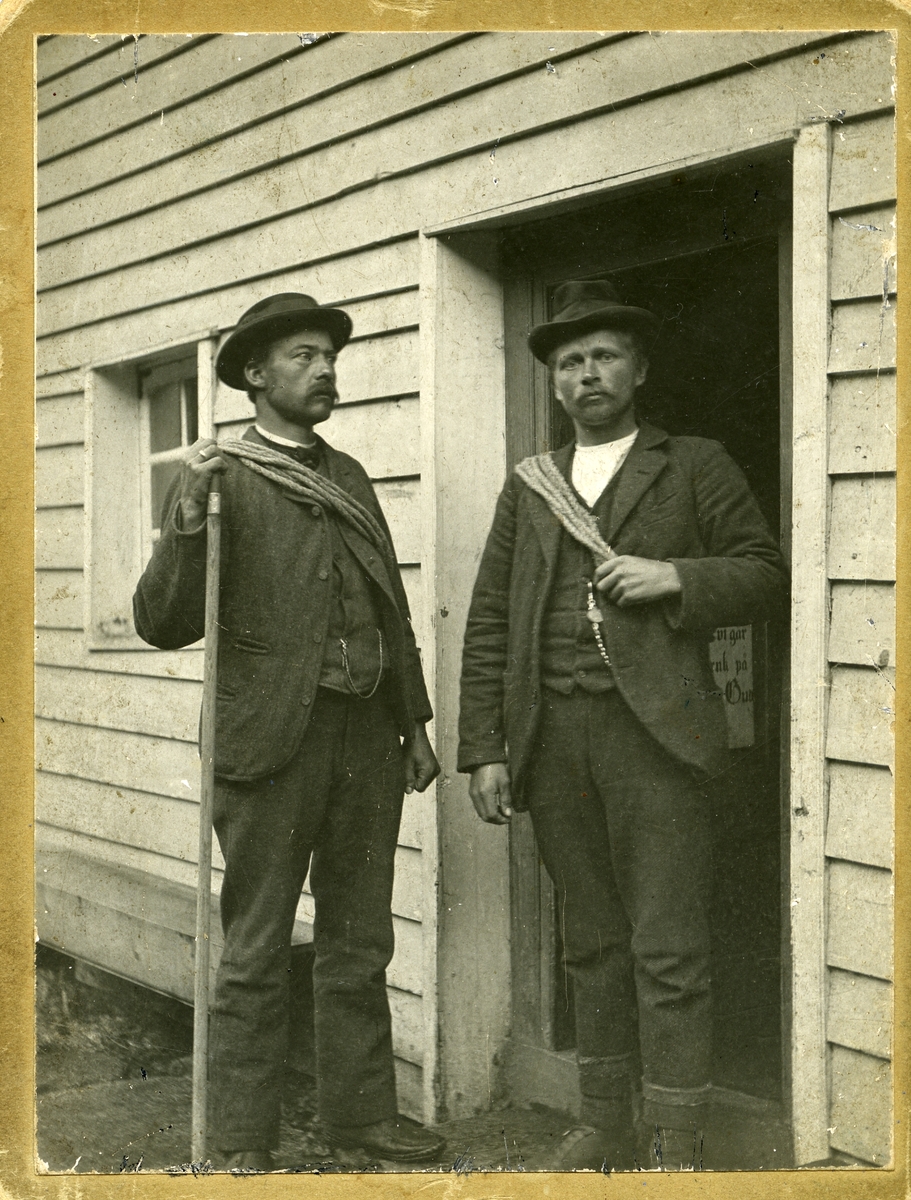 To menn foran en dør. Begge mennene har tau rundt overkroppen og en av dem holder en stokk i den ene hånden.