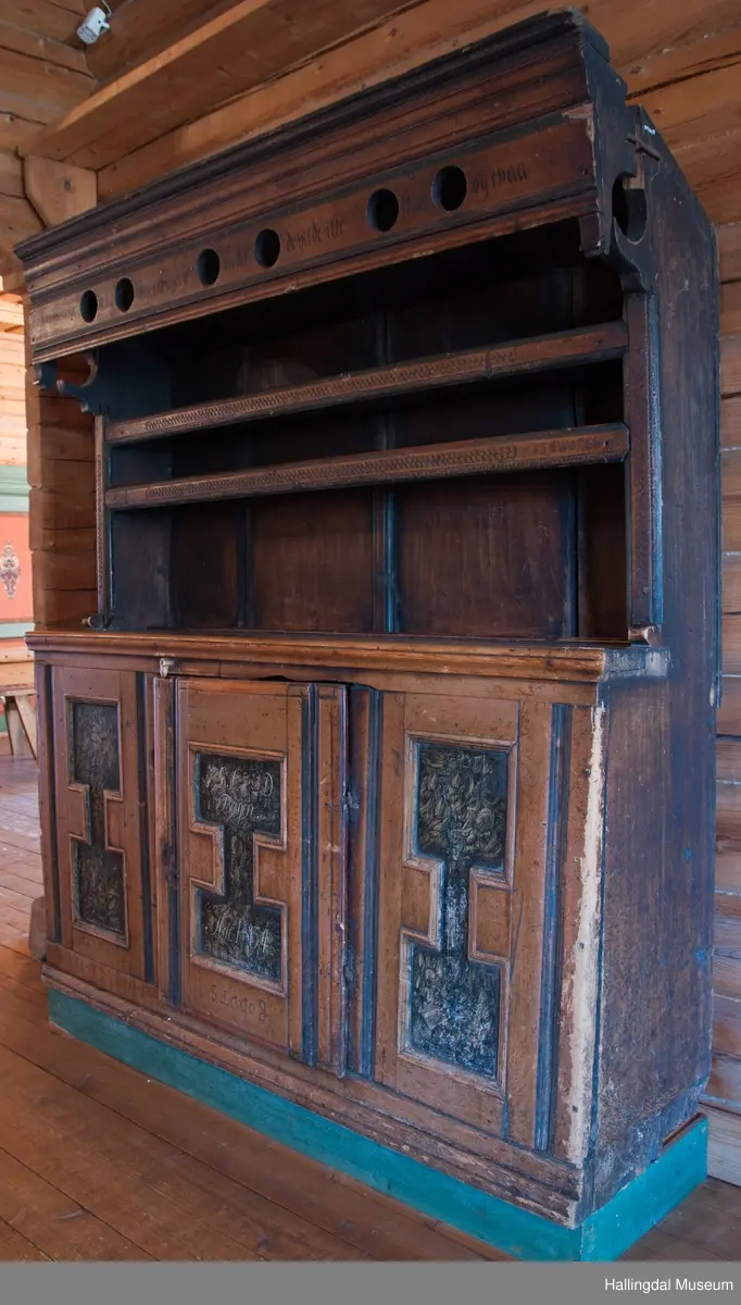 Rester etter rød og blå maling.  Gesims, skråner ned bakover. Profilert. 6 sirkulære hull i arkitraven.  Mellom disse er malt med sorte bokstaver.  Gavlene har utskårne "hylleknekter".  Bakveggen består av fire vertikale planker.  Skjøtene er dekket med profilerte lister.  Bare to biter i nederste del til venstre er fremdeles på plass.  Nederste del er lukket skap.  Tredelt, midterste del er skapdør.  Platen har profillist.  Feltene har speilfyllinger.  Blåmalte.  Klosset marmorering.  Speilfyllingenes form er som en liggen H.  Har profillist.  Skapet er satt på en ny grønmalt plate.  På døren er malt hvite bokstaver.  Nederst på døren er skåret inn bokstaver, opp-ned.  To nøkkelhull m. jernbeslag på venstre side av døren.  Over døren til venstre, en dreibar trekloss, holder igjen.  Innvendig har døren merke etter stort jernbeslag på samme side som nøkkelhullene.  To lister skåret i treverket minner om lange jernbeslag,-smalner mot nøkkelhullene.  Akantusranker i rødt, blått og hvitt, meget avslitt.  En hylle i skapet. Mangler bakvegg.  Karmen har på venstre side spor etter to jernhengsler.  Øverst på høyre kortside, mangler gesimsen en bit, og holdes til sideveggen med en jernkrampe.  Jernkrampe også på venstre side.  Skapplaten mangler list på høyre side.  Venstre side, fra gesimsen og ned har spor etter å ha stått inntil en vegg eller et annet møbel.