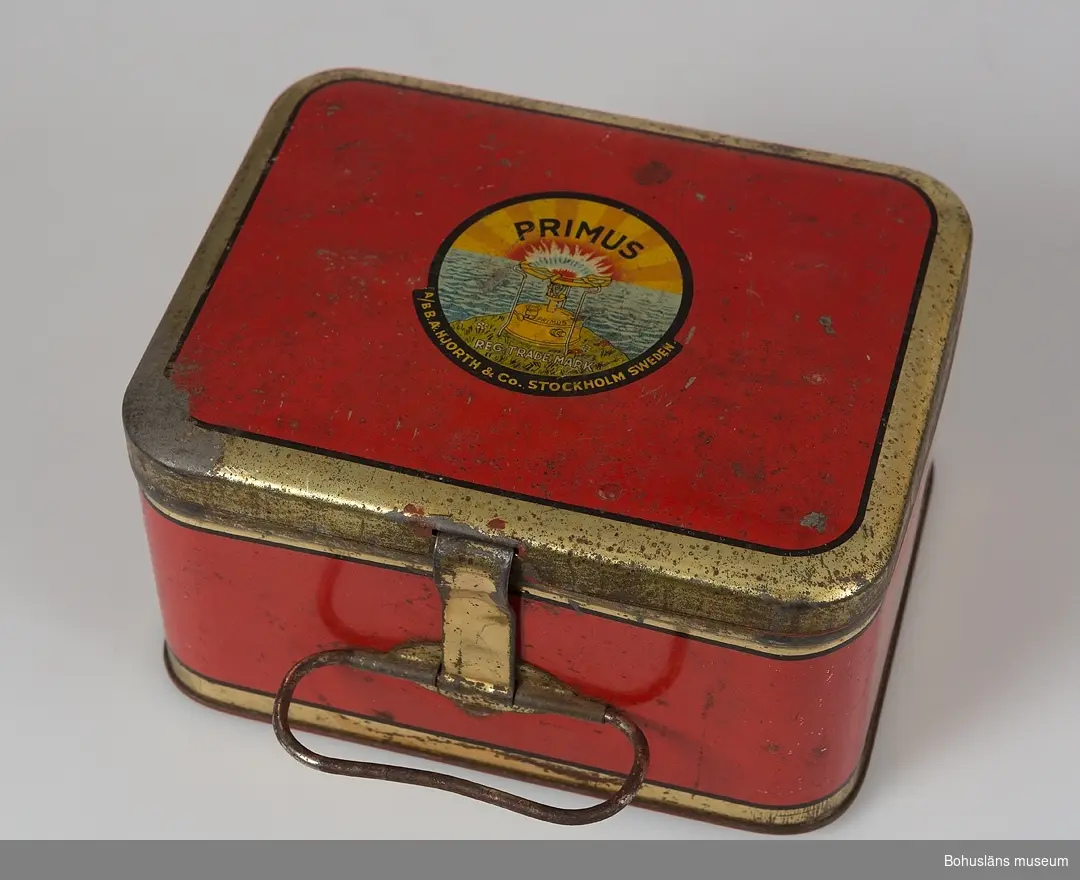 Primuskök med liten bränsledunk och nyckel och låda i metallplåt att förvara sakerna i. Lådan i röd färg och text "Primus" på locket.