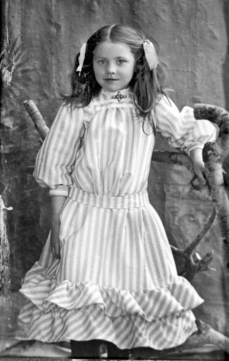 Kjersti H. Sisjord som jente, ståande i fotoatelier, fotografert kring 1908.