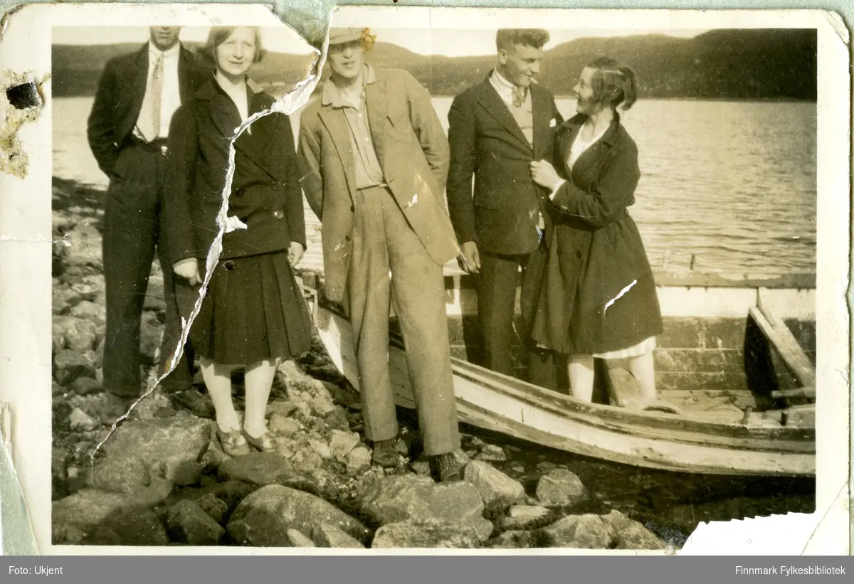 Gruppeportrett av kvinner og menn. De fleste på bildet er ukjente, men mannen som står i båten er Petter Aule. Mennene er kledd i jakker, bukser, slips og hatter. Kvinnene i kjoler, strømpebukser, sko og jakker. Gruppen står på steiner. Bildet er tatt i Jarfjordbotn. De andre avbildete personene kan være fra Vardø.