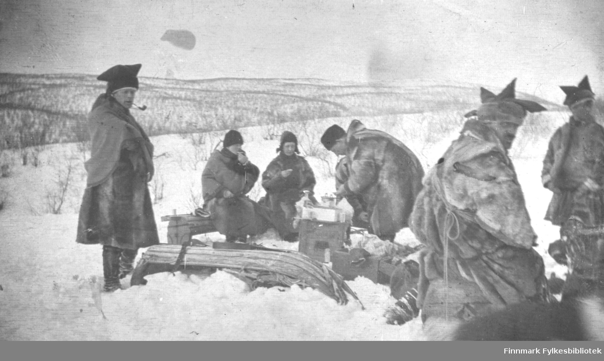 Aimjok,1916. Samer i pesk med karakteristisk samisk lue. En av mennene røyker pipe. Noen sitter med mat eller drikke i hendene.