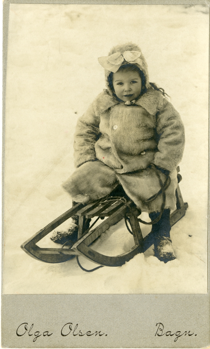 En liten pike avbildet sittende på en kjelke. Jenta er iført varm vinterjakke og lue med sløyfe på.