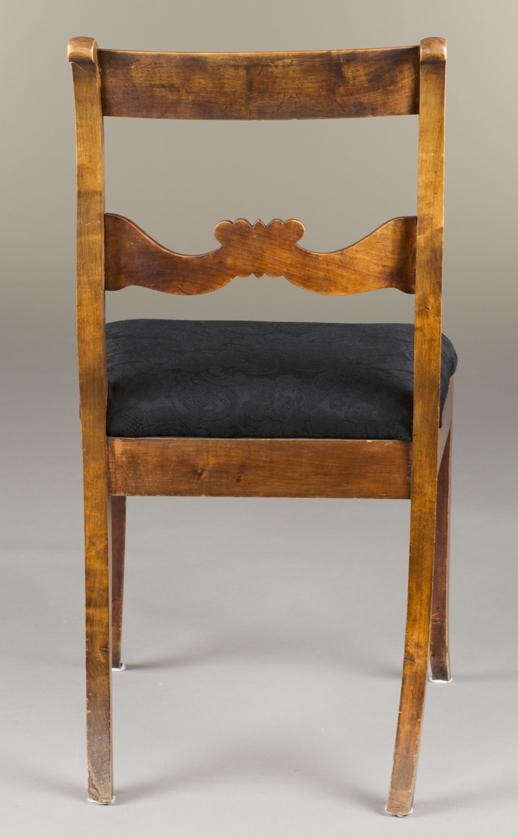 Biedermeier-stol med svart trekk. Stolen er av tre og har fire ben. Polert treverk. Alle bena er svungne. Utskjæringer på midtstykket på ryggen. Svunget og avrundet toppstykke på rygg. Polstret og trukket med et svart vevd materiale.