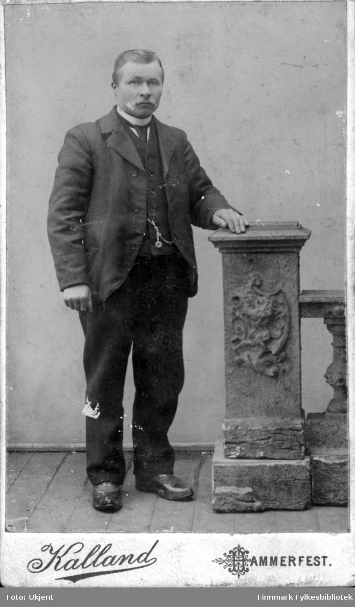 Et portrett av Peder Isak Mathisen (f.1872) som arbeidet som handelsmann i Nuvsvåg. Han er kledd i dress: jakke, vest, slips, bukse og sko. På vesten henger kjedet til et lommeur. Han hviler hånden på en søyle.