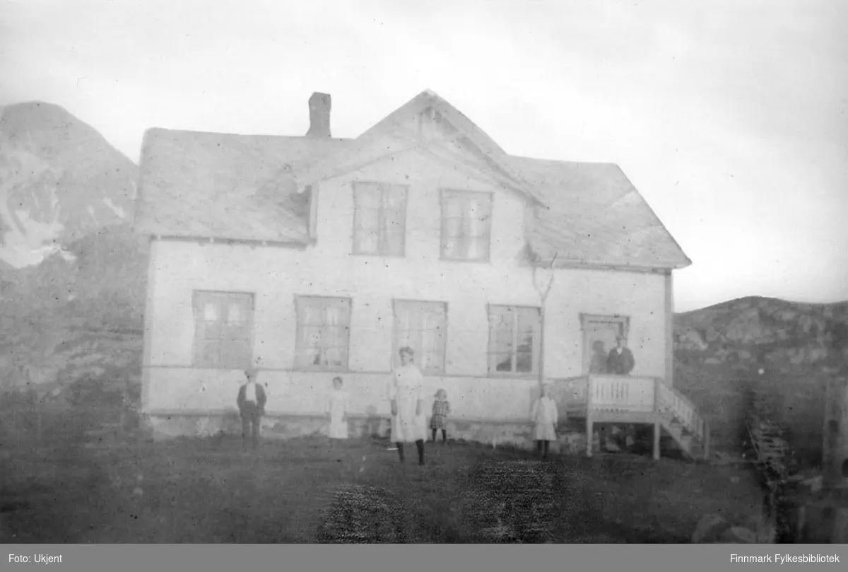 Peder Mathisens handel og bolighus på Skolten, Nuvsvåg. Bildet tatt omkring 1920. Peders barn er samlet utenfor huset.  Man kan se et hus med vindu, trapp, pipe, grunnmur og et tak.