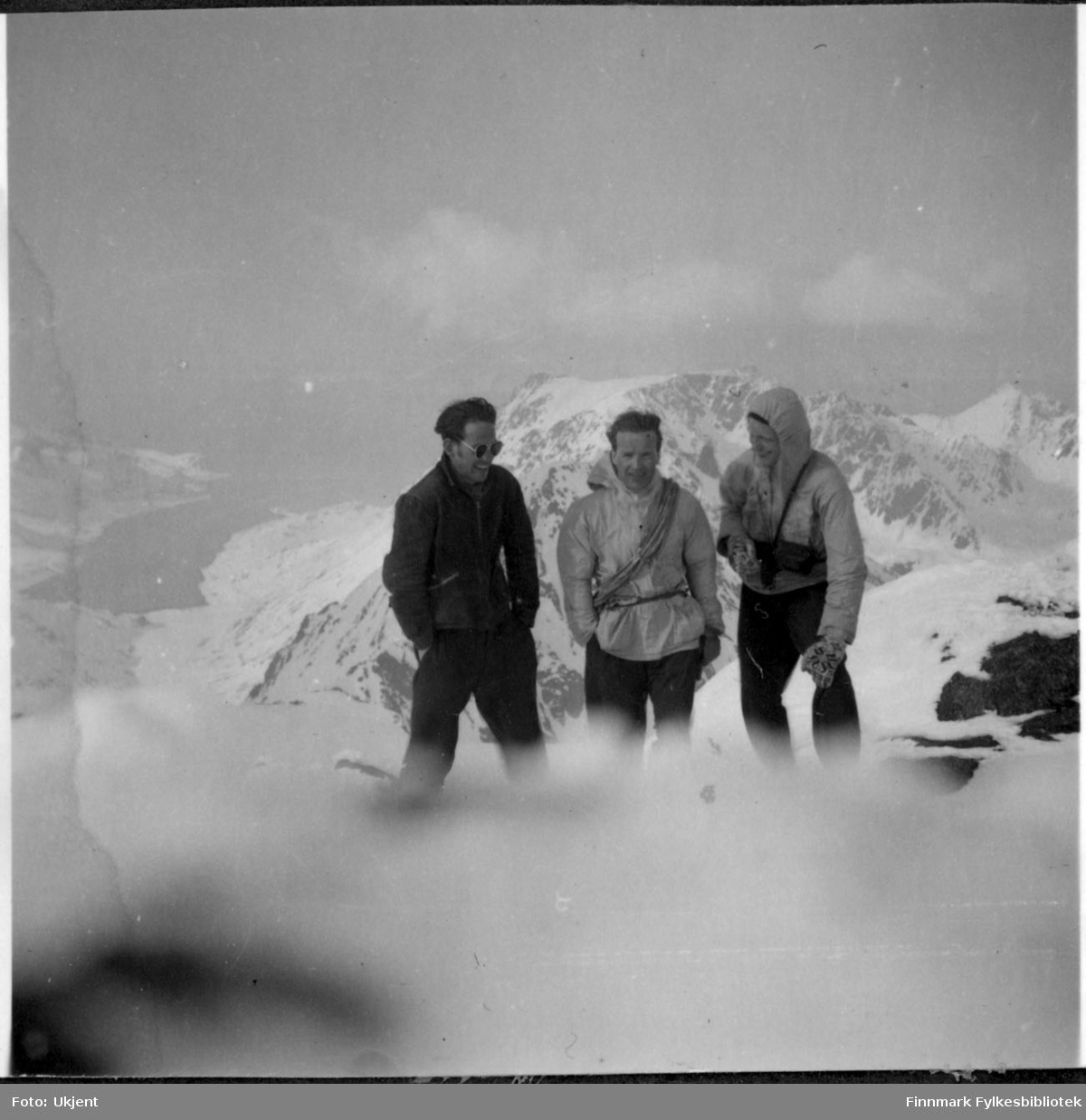 På tur til Øksfjordjøkelen i Nuvsvåg. Fra venstre Gunnar Ørstadvik, Adolf Blone og Reidar Martinsen. De tre mennene har på seg vinterklær, jakker og bukser. Gunnar helt til venstre har på seg solbriller og man kan se at Reidar helt til høyre har på seg votter. Bak mennene kan man se fjell og vann. Det er mye sne på bakken.