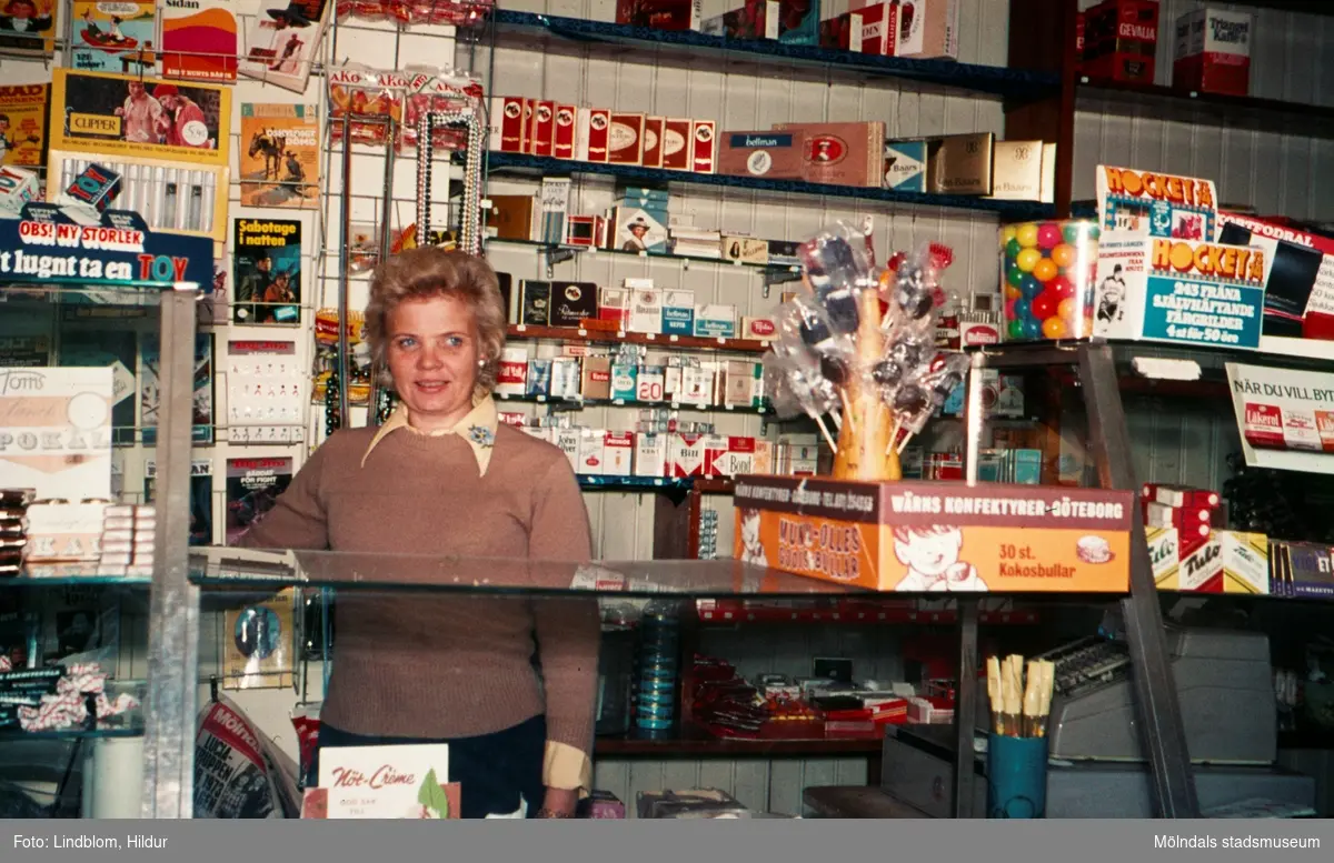 En kvinna bakom disken i en tobaksaffär, år 1973. Möjligen vid eller i närheten av Gamla Torget i Mölndal.

För mer information om bilden se under tilläggsinformation.