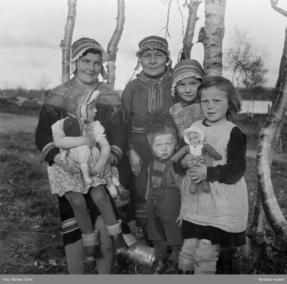 Porträtt av tre generationer samiska kvinnor och barn. Flickorna med dockor i famnen. Britta Prost (född Sevä 1912) med dottern Elin Ragnhild (född 1944), svärmor/faster Susanna Prost (född Sevä) och brorsbarnen Ellen Margareta och Karl Erling. Från Nordiska museets Lappmarksundersökning.