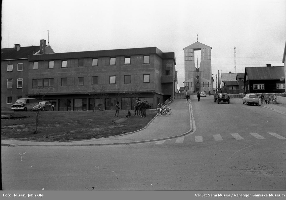 Gatebilde fra Vadsø sentrum, hotellbygningen til venstre og Vadsø kirke øverst i bildet. En traktor med blant annet melkespann på hengeren er på vei opp bakken. 1967.