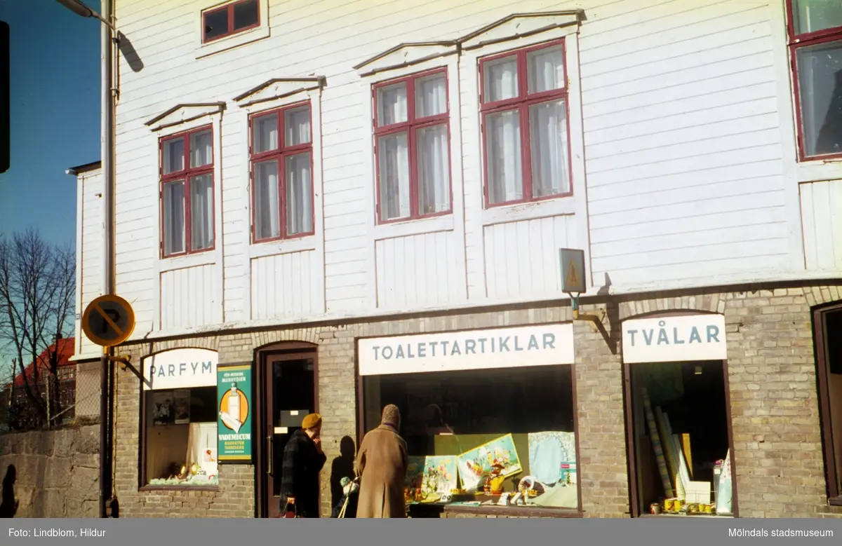 Två damer tittar in genom skyltfönster till kemikalieaffären Stella med adress Kvarnbygatan 23, Eneborg, i Mölndal, 1960-tal.

För mer information om bilden se under tilläggsinformation.