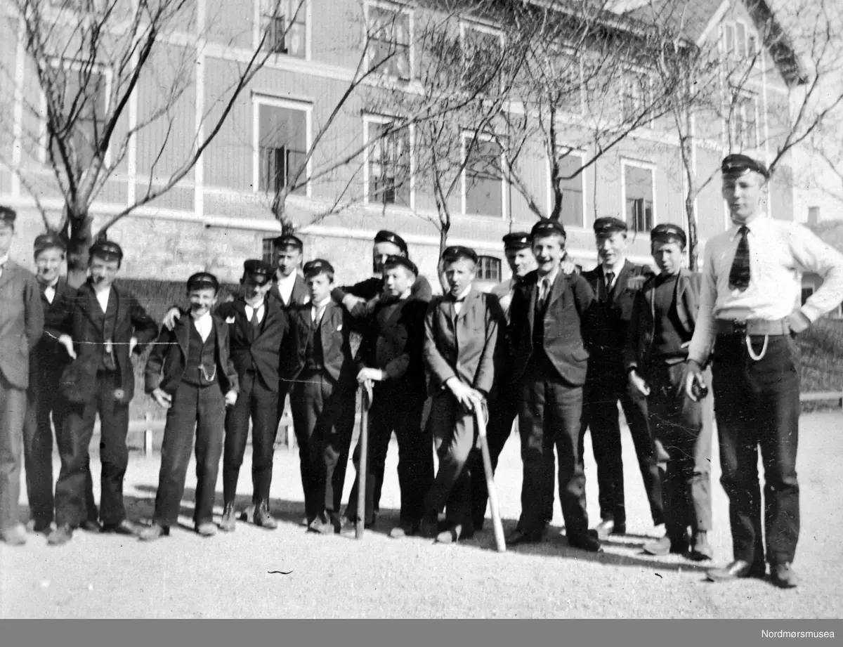 "En gruppe middelskolegutter ved skolen, antakelig 1902 eller 1903. Jeg drar kjennsel på bare to: "x" fra venstre: Håkon Olsen, nå tannlege i Haugesund (med stokk). "X" Einar Trumpy, nå øyenlege i Oslo" (Kommentar: Olaf Yderstad kom til skolen som adjunkt fra 1. august 1903). Fra Nordmøre Museums fotosamlinger. Artianere?
