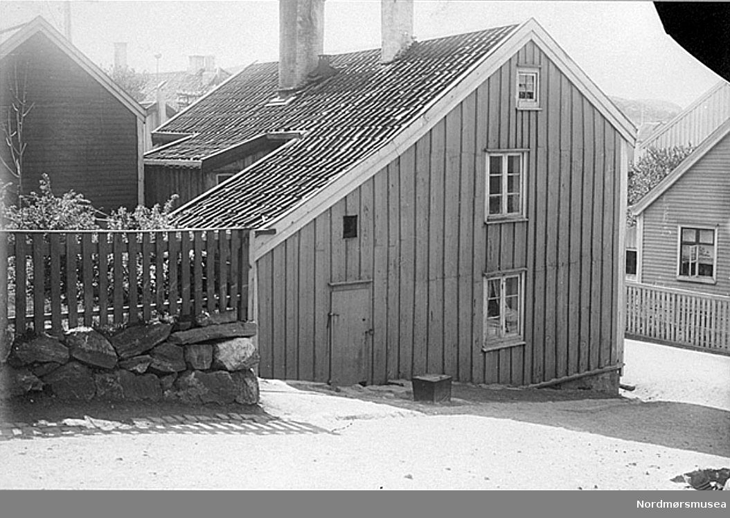 Arbeiderbolig for firmaet Parelius & Lossius, eid av  Rasmus Brochmann Parelius (1821-1901) og seinere av hans sønnesønn Rasmus B. Krogh. 
Huset har to etasjer. Tre hybler og felles kjøkken (med skorstein) i første etasje. Det har tømmermanns-bordkledning som delvis er rødmalt, delvis gulmalt.  Taket er belagt med røde teglstein og har takrenner av tre, noe skadet.  På baksiden av huset er et utbygg og et påbygd karnapp på pæler –for lager/uthus. På bildet ser vi utbygget med egen inngang i forgrunnen;  karnappet er lengre bak.  Husets forside med hoved-inngangsdøren vender ut mot gaten som sees til høyre på bildet.  På nedsiden mot gaten har huset en lav grunnmur.  På baksiden, som ligger lengre oppe i bakken, sees ingen grunnmur.  En leieboer i huset opplyser at huset ikke har kjeller.  Et lite klosett (utedo) står bak huset.  I gangen er det tømmervegg.  I kjøkkenets skorstein står en liten kokeovn.  Leieboeren i denne etasje av huset beretter at man på denne ovnen koker over åpen ild på brannfot ved klesvask, mens der ellers kokes i den i ovnen/skorsteinen stående lille kokeovn. 
En brannfot er et lavt stativ av jern som takke, stekepanne eller kokekar kan settes på når varmekilden er åpen ild i grua. Brannfoten er vanligvis trekantet eller rund med tre føtter. De fleste er smidd av lokale smeder, men det var også jernverk som støpte slike produkter. Gryter og kjeler med hank eller ører ble hengt opp i skjerdinger, brannfoten ble først og fremst brukt til stekeredskaper uten oppheng.
I gaten utenfor husets ene gavlvegg står en vask av tre.  Denne sees på bildet.  Ifølge en av husets leieboere brukes kun denne vask av hele husets beboere, og ifølge samme finnes der ingen vask inne i huset.  Huset eies ifølge samme av firmaet Parelius og Lossius (Krogh) og beboes av arbeidere i dette firmas tjeneste. -- 


-- Katalog over bilder av eldre gateparti, bygninger og bygningsdetaljer i Kristiansund.
Bildene er bekostet av Det Kulturhistoriske Lag (DKL)og med bidrag av Kristiansund Sparebank og fru Caroline Knudtzon.   Ført i protokoll av overlærer Wilhelm Lund.  Foto hovedsakelig utført av fotograf  Ole Olsen Ranheimsæter dersom ikke noe annet er anført.  Tegninger utført av billedhugger J. Johnsen og Ole Ranheimsæter. 
DKL -  er en forløper for Nordmøre Historielag, i tett samarbeid med Kristiansunds museum.
Nr. 113  Hus paa Nordlandet, Kristiansund, lnr. –
Huset er 2-etages, og planen av første etage er som paa hosstaaende rids x)

Skisse:               

A = gang
B = kjøkken med skorsten ( c )
D = værelse
E = værelse
F = værelse
G = utbygg
I = gatedør
H = karnap.

Huset er et mindre hus.  Det har fømmermandsbordklædning som delvis er rødmalet, delvis gulmalet.  Taket er belagt med røde teglstensheller.  Langs takkanten gaar ialfald for en del vandrender av træ, som ligger op paa kanten av nedtaket, hvis nedre ender sees under renden, og hvis underliggere er haandskåret.  På baksiden av huset er et utbygg (G i planen) og et utbygget karnap ( H i planen).  Dette karnap hviler paa stætter og naar altsaa ikke ned til jorden,  hvad utbygget G derimot gjør.  Paa billedet sees dette utbygg i forgrunden;  karnappet sees længere bak.  Husets forside med indgangsdøren vender ut mot gaten som sees til høire paa billedet.  Paa nedsiden mot gaten har huset en lav grundmur.  Paa baksiden, som ligger længere oppe i bakken, sees ingen grundmur.  En leieboer i huset oplyser at huset ikke har nogen kjelder.  Utbygget ( G ) er ialfald delvis av panel og brukes til uthus.  Karnappet ( H ) er, saavidt sees, av panel og brukes vist til uthus.  En av husets leieboere oplyser at der til huset ikke hører andre uthus end utbygget (G) og karnappet ( H ) samt et liltet kloset, som staar bak huset.  I gangen ( A ) sees tømmervæg.  I kjøkkenets skorsten (paa planen C ) staar nu en liten kokovn.  Leieboeren i denne etage av huset beretter at man endnu paa denne skorsten koker over aapen ild paa brandfot ved klævask, mens der ellers kokes i den i skorstenen staaende lille kokovn.  I gaten utenfor husets ene gavlvæg staar en vask av træ.  Denne sees paa billedet.  Ifølge meddelelse av en av husets leieboere brukes kun denne vask av hele husets beboere, og ifølge samme findes der ingen vask inde i huset.  Huset eies ifølge samme av firmaet Parelius og Lossius (Krogh) og beboes av arbeidere i dette firmas tjeneste

Fra Nordmøre Museum sin fotosamling.