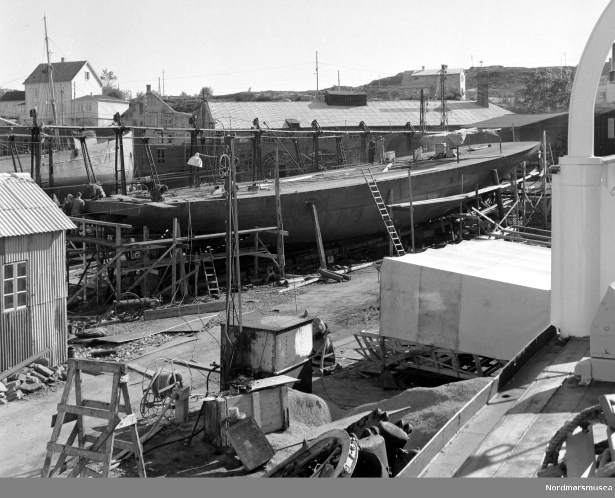 Bildet viser B/F";Norddalsfjord"; bnr.14 ved Storviks Mek. Verksted under bygging på beddingen. "Norddalsfjord" ble levert til Møre og Romsdal Fylkesbåtar 15. mars 1961 og hadde følgende hoveddimensjoner: L 31,20 m x B 8,55 m x D 3,35 m og hadde en tonnasje på 159 bruttoregistertonn. Fremdriftsmaskineriet består av 3 Volvo Penta turboladede dieselmotorer type TMD96 på til sammen 420 hk som via kilremdrift var koblet til et felles gir og propellaksel med vribar propell, slik at hver enkelt av motorene kunne kjøres separat. Fergen hadde 2 Bolinders vekselstrøms-aggregater type 1052MG på 23 hk hver tilkoblet en generator på 17 kW. Fergen var utstyrt med elektrohydraulisk styremaskin. Fergen har plass til 18 personbiler og har sertifikat for 160 passasjerer. Forut er det innredet 6 lugarer for offiserer og restaurantpersonale og akterut en mannskapslugar for 4 personer og toppfarten er 11,4 knop og marsjfarten 10,5 knop. Ferga er verkstedets første nybygg etter B/F"Trygge" som ble levert i 1938.

Bak nybygget ses gangveiene på patentslippvogna klinkbygd i stål 1918, den største slippen mellom Bergen og Trondheim. Det lange bygget i bakgrunnen med grått tak er kjelverkstedet fra 1905 og revet etter 1980. 

Til venstre for dette ses den blå kontorbyggningen, bygget ca. 1902 og revet og brent på 1970-tallet, og øverst, til venstre, et av bolighusene til verkstedet, bygget opp av et gammelt meieribygg fra Straumsnesset ca.1910 og brant ned på 1980-tallet, hvor familien Hjelmås bodde, senere familien Paul Storvik. I bolighuset, til høyre, over taket på kjelverkstedet, bodde familien Lisbeth Kvalnesvik, familien Magnus Kvalnesvik og familien Ludvig Bergem. Til høyre for det, bak daviten ses huset til familiene Alf og Knut Hjelmås. Foran nybygget ses blikktaket over innkjørselen til verkstedet. Det står en båt på såpehellingen fra 1901, sansynligvis M/S"Greip". Båten i forgrunnen, som vi ser livbåtdekket og daviten på, er Ole T. Flakkes lastebåt M/S"Knoll" som ligger ved piren. Til høyre på bildet, foran nybygget, ses et presenningsoverbygg over sveiseplanet. Bygget helt til venstre i bildet inneholder verkstedets dampkjel og det lille huset, på piren, mellom dampkjelhuset og presenningsoverbygget er for strømuttak og trykkluftuttak. Bildet er fra 1960. Samme som FAKf-100296.214953. Fra Nordmøre museums fotosamlinger.



