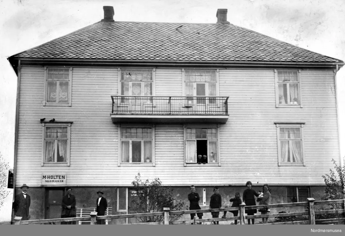 Diverse personer i forgrunnen av Lars Guttormsens gate 21 på Kirkelandet. Merk skiltet ";M. Holten skomaker";. balkong Bildet er datert 1932. Fra Nordmøre Museums fotosamlinger
