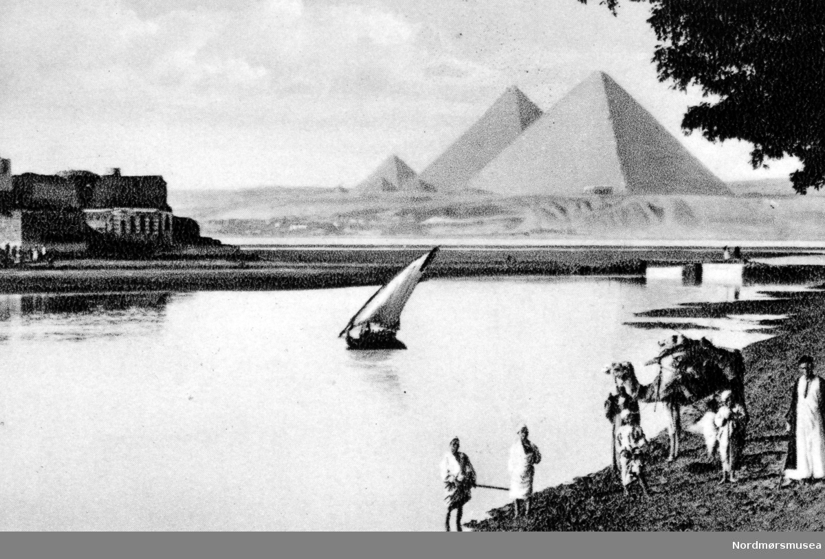 Postkort fra Egypt, hvor vi ser en gruppe mennesker med kamel nede ved elven, og Giza pyramidene på den andre siden. Fra Nordmøre museums fotosamlinger. /Reg:EFR2013/



