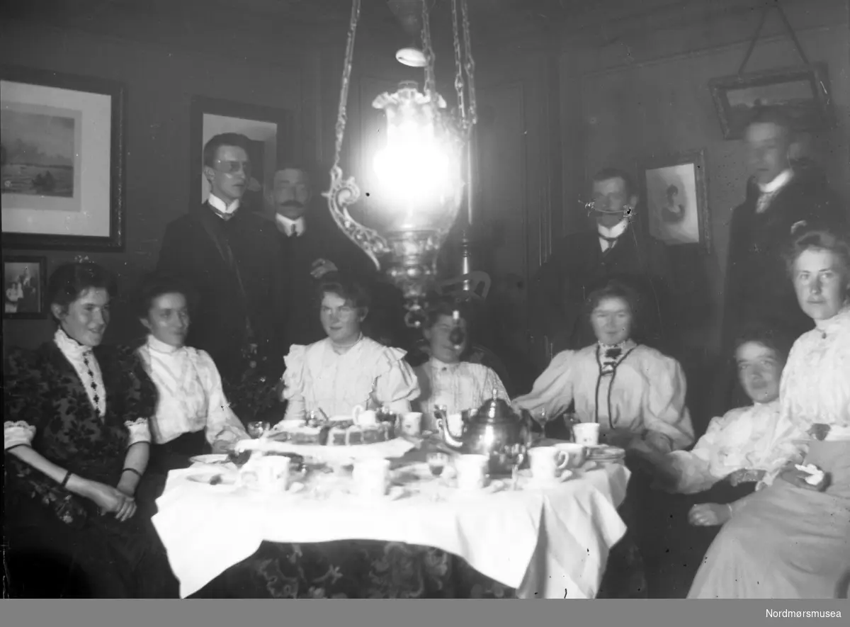 Foto fra et teselskap, hvor vi ser syv kvinner sittende rundt et bord mens fire menn står like ved. Det er ukjent hvem vi ser på bildet, samt hvor og når bildet er tatt. Fra Nordmøre museums fotosamlinger. Reg: EFR
