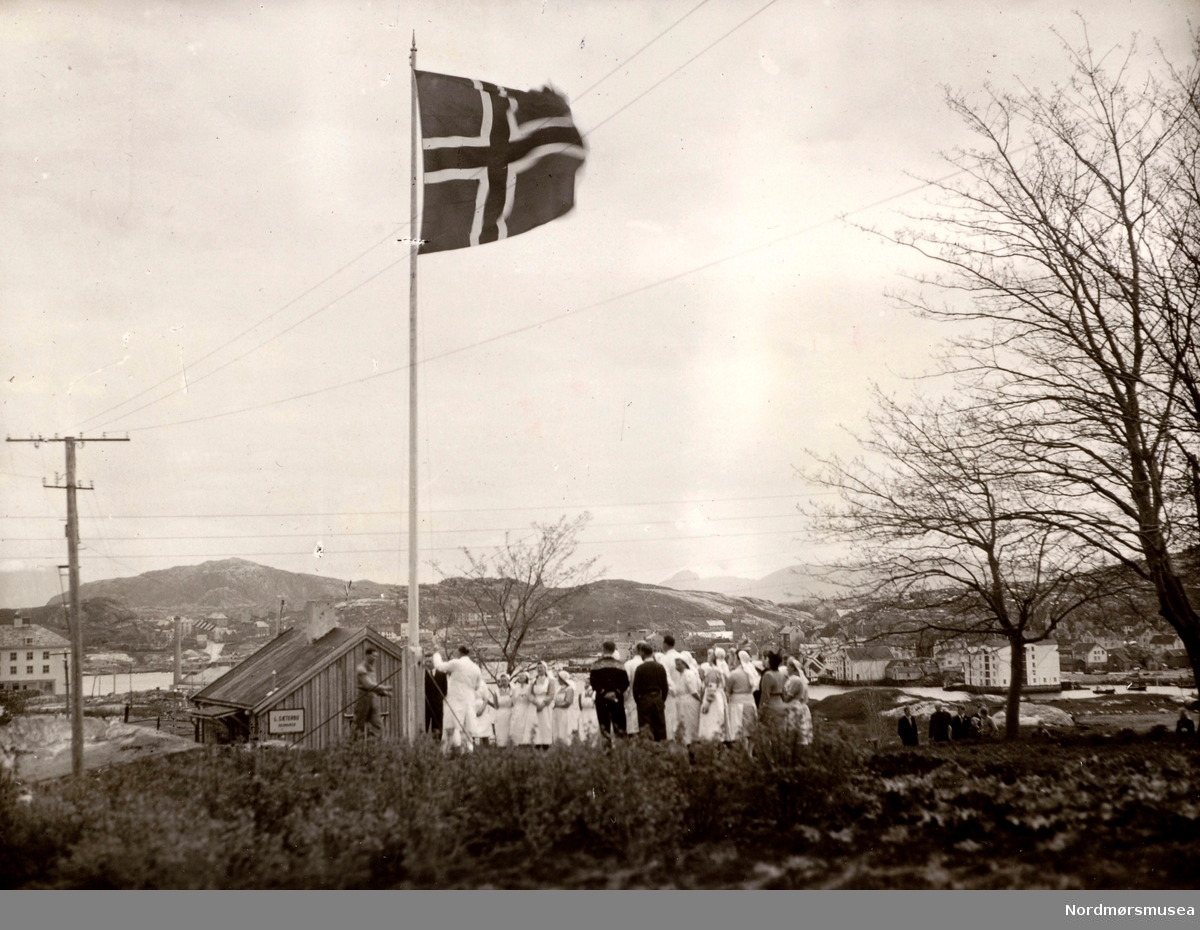 Foto fra 8. mai 1945, hvor vi ser en gruppe sykesøstre heise det norske flagget på frigjøringsdagen. Bildet er fra Kristiansund. Syepleierstudentene var fra Røde Kors' søsterhjem. Fra Ida M. Knudtzons fotosamlinger, nå en del av Nordmøre Museums fotosamlinger.