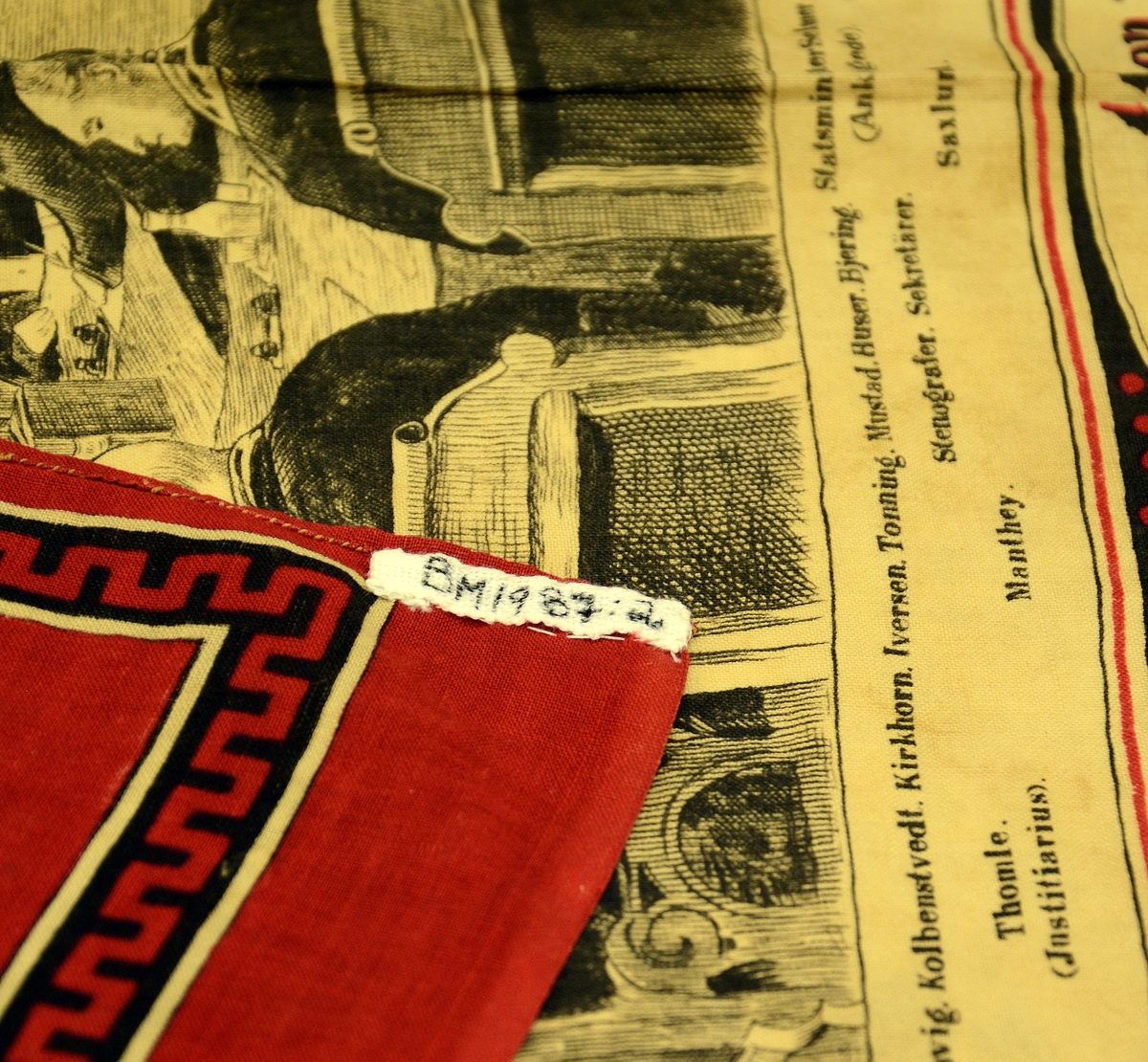 Tørkle med påtrykt motiv "Rigsretten 1883". Bilde av riksretten samlet i rettssalen med navn på personene under. Rundt bildet en bord på rød bunn. Tørkledet har flere hull. Håndsøm rundt to av sidene.