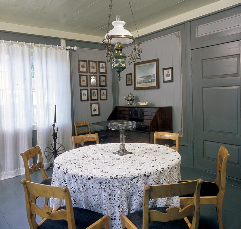 Interiør; rundt bord med hvit duk og stoler rundt i forgrunnen, husordel, mange fotografier på veggen og en oljelampe over bordet. (Foto/Photo)