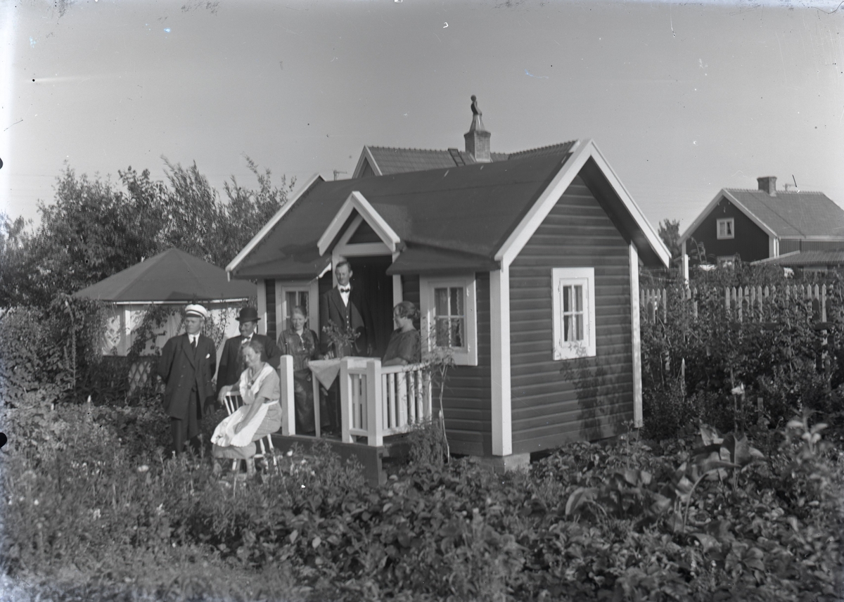 En stuga, tre män och tre kvinnor i Kalmar södra koloniområde, fotograferat omkring 1930. Kalmar södra koloniförening grundades 1917 och har idag 105 kolonilotter. Området ligger strax söder om länssjukhuset i Kalmar med huvudingång från Stensbergsvägen.