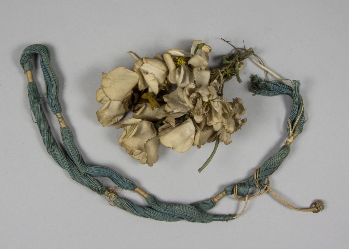 Blombukett, rosor, tillverkad av tyg och överklädda metalltrådar. Hopbundet med blombuketten en härva blått bomullsgarn omknuten på flera ställen. Från härvan hänger en liten mässingsknapp.