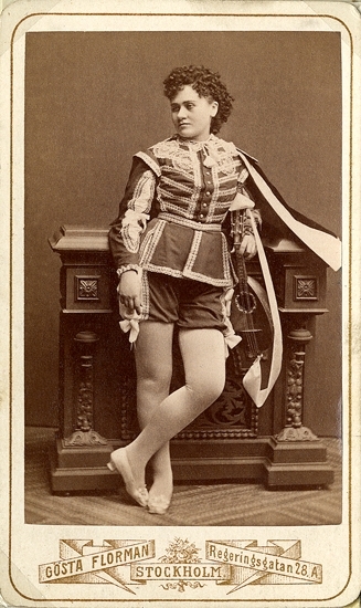 Rollporträtt (helfigur) av operasångerskan Carolina Östberg i mansdräkt med cape.Carolina Östberg -  1853-1929, var en svensk operasångerska.Hon var dotter till en fältskär och visade tidigt musikalisk begåvning, som hon utvecklade på Dramatens elevskola 1869-1873. Hon var elev till Signe Hebbe. Hon visade sin talang som konsertsångare innan hon debuterade på Stora Teatern 1873 tillsammans med Louise Pyk med varsin roll i samma pjäs. De gjorde båda succé; Östbergs röst ansågs som en frisk klar och ren timbre, en svag men ren sopran med bra koloraturisk träning, och hon hälsades med jubel av publiken i mansrollen Carlo Broschi i "Hälften var" 1874, varefter hon fick fast anställning; rollen som Carlo Broschi förblev hennes mest populära, och fram till hennes kontrakt utgick 1877 gjorde hon en rad stora roller och utvecklades mer för varje gång.Då kontraktet gick ut 1877 tänkte hon resa till Ryssland, men äktenskapet med grosshandlaren Horwitz 1878 fick henne att avsluta sin yrkesverksamhet, som det troddes för alltid. Hennes avgång ansågs vara en stor förlust; på kort tid hade hon blivit landets kanske mest populära operasångare.Då Carolina Östberg-Horwitz oväntat annonserades som gästskådespelare i titelrollen "Boccaccio" på Nya Teatern 1879 drog förställningen fulla hus, och hon hälsades med så stor entusiasm att "jublet aldrig ville ta slut"; hon gjorde en stormande succé, kallades hjältinna och visade talang både i lättsamma operetter och mer allvarlig opera, och pressen uppmanade Operan att anställa henne. Hon turnerade i Köpenhamn 1880 och Tyskland 1882-1883 , Norge 1885 och tillhörde Nya Teatern fram till 1886. Hon använde sitt ursprungliga efternamn kallade sig själv fru Östeberg.År 1886 anställdes Östberg igen på Operan, vilket sågs som en triumf för publiken; "publiken hade fått sin vilja fram" mot Operan, efter att i flera år krävt att hon skulle anställas där. Hon stannade sedan där, förutom en turné in USA 1892-94. Hon jämfördes med Louise Michaëli. Hon fick Litteris et Artibus in 1891, med briljanter 1900, och invaldes i Musikaliska akademien 1897. 1905 gjorde hon 9 grammofoninspelningar, och 1906 slutade hon sjunga och var sedan sångpedagog.Bland rollerna fanns Gioconda, Sieglinde i Valkyrian, Alice i Falstaff, Margareta av Valois i Hugenotterna och Aida.