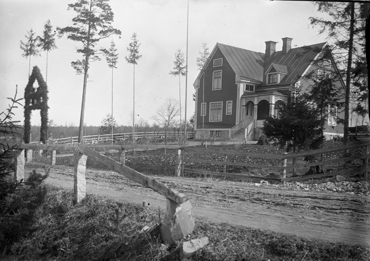 Ett bostadshus med en inte riktigt färdig trädgård. Vid ingången syns en bröllopsportal smyckad med granris och ett antal uppställda höga granstörar.
Trol. Fritz och Agda Bergqvists första hem, 1918.