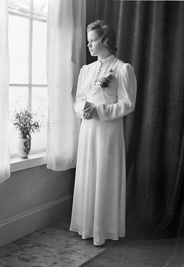 Foto av en kvinnlig konfirmand i lång, vit konfirmationsklänning som står vid ett fönster. Hon håller en liten psalmbok i händerna.
Helfigur. Ateljéfoto.