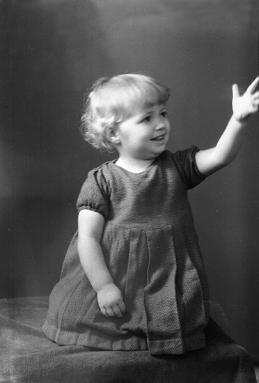 Foto av en liten flicka i mörk ylleklänning, som står på knä på en sammetsduk.
Helfigur. Ateljéfoto.
Saida Fick-Blomberg (1933-1998), Skärholmen. F. i Aringsås.
Källa: Sveriges Dödbok 1901-2009.