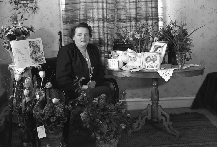 Foto av en medelålders kvinna som sitter i ett vardagsrum, omgiven av mängder av blommor och presenter.