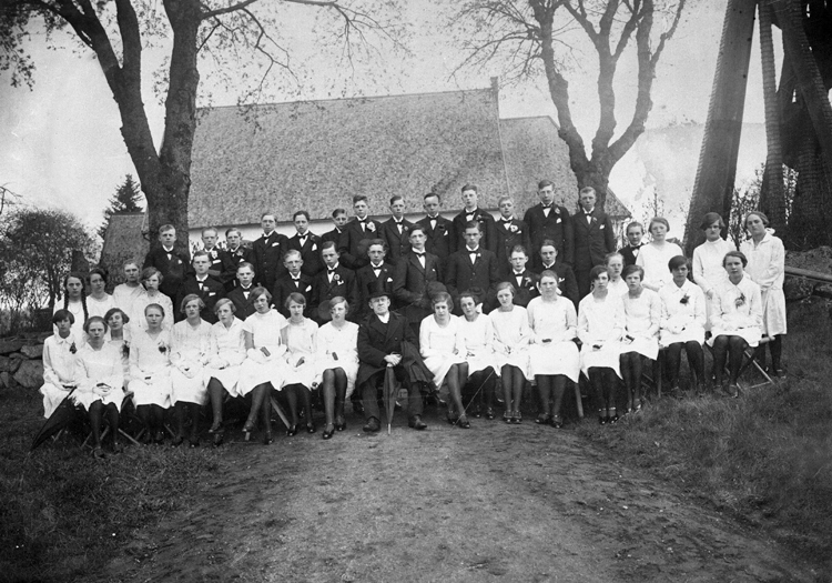 Foto av en grupp konfirmander utanför Aringsås (Alvesta) kyrka tillsammans med deras läspräst, kyrkoherde Ivar Björnberg (1861-1933), kyrkoherde i Aringsås och Lekaryds församlingar från 1908.