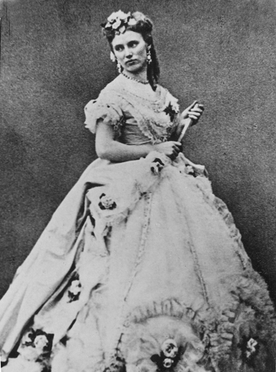 Porträttfoto (rollporträtt ?) av Christina Nilsson. Hon bär en stor klänning (krinolin) med blommor på. I handen håller hon en solfjäder. Hon bär även halsband och örhänge. Violetta i La Traviata.