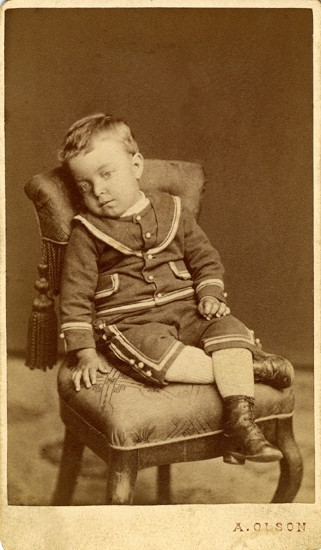 Porträtt (helfigur) av en okänd pojke i mörka knäbyxor och dito jacka, prydda med ljusare garneringar. 
Han sitter på en stoppad stol, prydd med en tofs på ryggstödet.