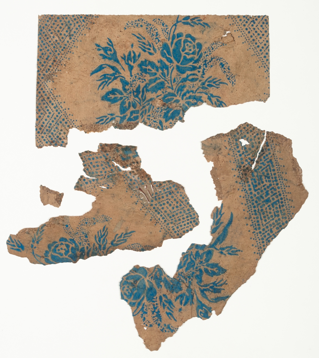 Fragment av tapet med rombmönster med mittmotiv i form av en rosenbukett i blått på obestruken botten. En tryckfärg. Har varit tapetserad över 28741. IB