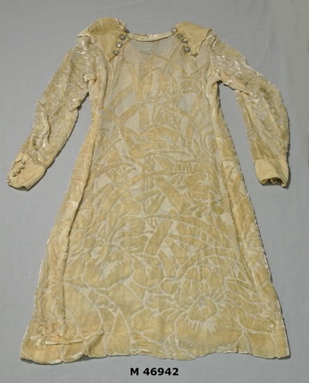 Cremefärgad klänning i crepé de chin med utskuren silkessammet.  
Krage draperad fram, fästes bak med 8 ( 4 på var skuldra ) glas/metallknappar. Klänningen rakskuren med snäv ärm, manchett med 5 st klädda knappar.
Ärmläng  480 mm.

Inskrivet i huvudbok  1985