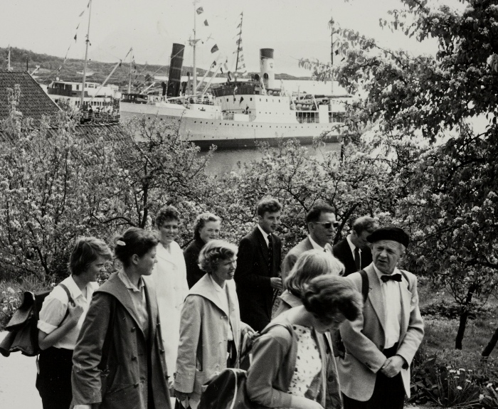 Mennesker på spasertur fra kaien på Nedstrand under sommerturen til Jonas Øglænd Sandnes 08.06.1958. I bakgrunnen ligger båtene på kaien