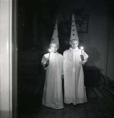 Med strut på huvudet och ljus i hand lussar två pojkar den 13 december 1951 i Glössbo. Pojkarna står i ett mörkt rum klädda som stjärngossar i vita lucialinnen.