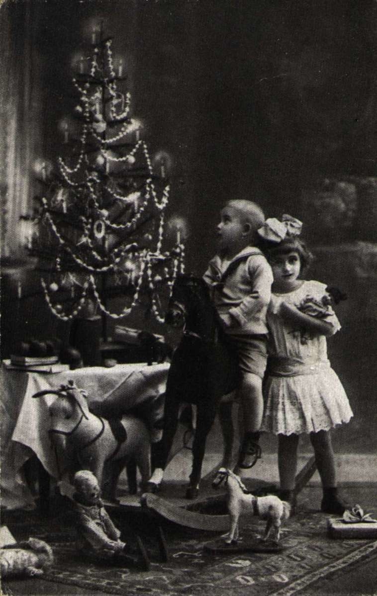 Julekort. Jule- og nyttårshilsen. Svart/hvitt fotografi. Gutt og pike foran pyntet juletre på bord. Julegavene i forgrunnen.
