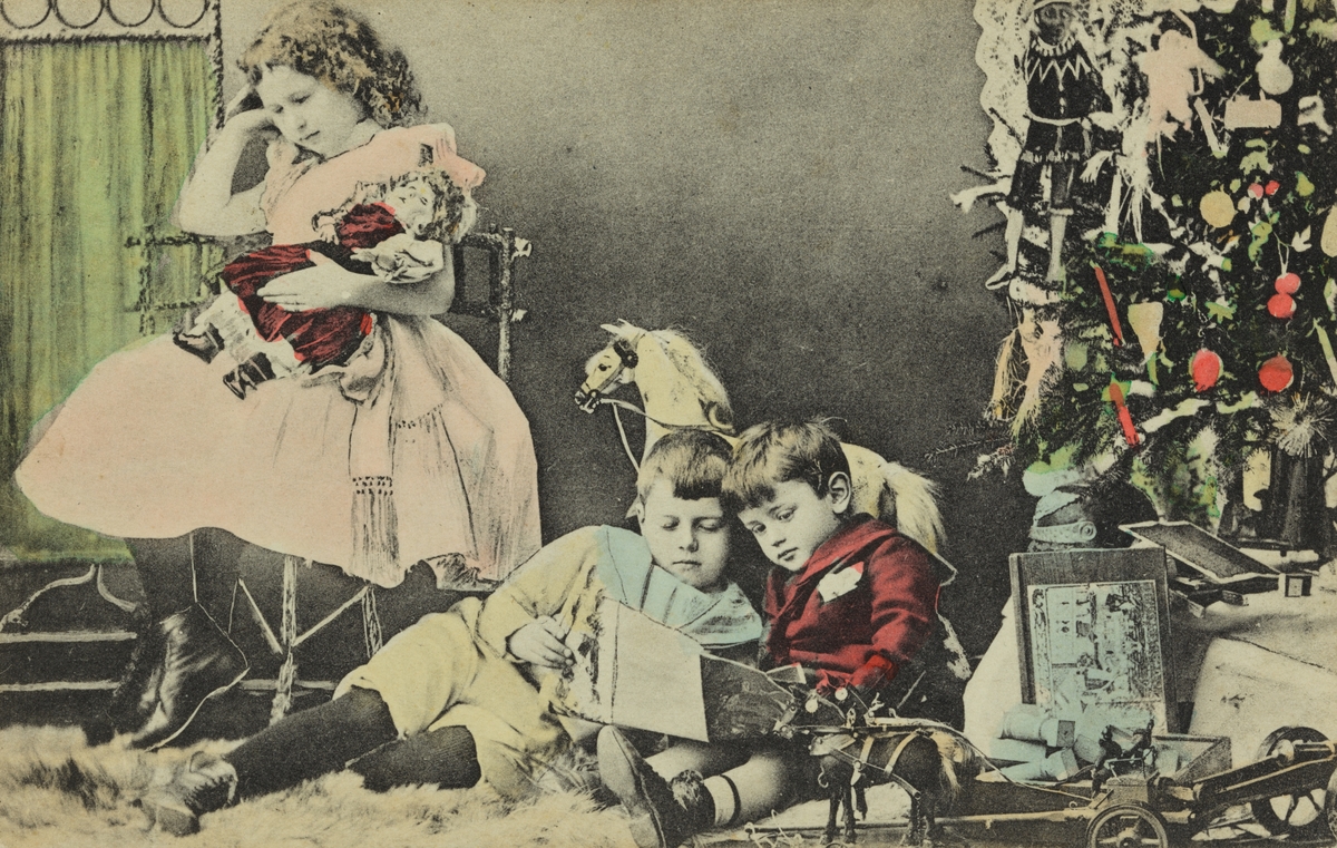Julekort. Jule- og nyttårshilen. En pike og to gutter med julegaver foran et pyntet juletre. Håndkolorert fotografi. Stemplet Larvik 23.12.2011.