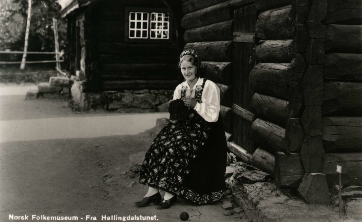 Postkort. Kvinne i bunad sitter i husveggen og strikker. Hallingdalstunet, NF.