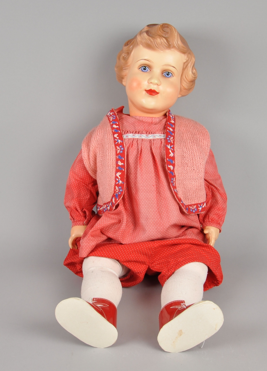 Dukke av celluloid. Dukken gikk under navnet "Gro-dukken). Dukken har påmalte øyne, munn, hår og kinn. Den er kledd i kjole, bukse, strømper, vest, sko og undertøy.