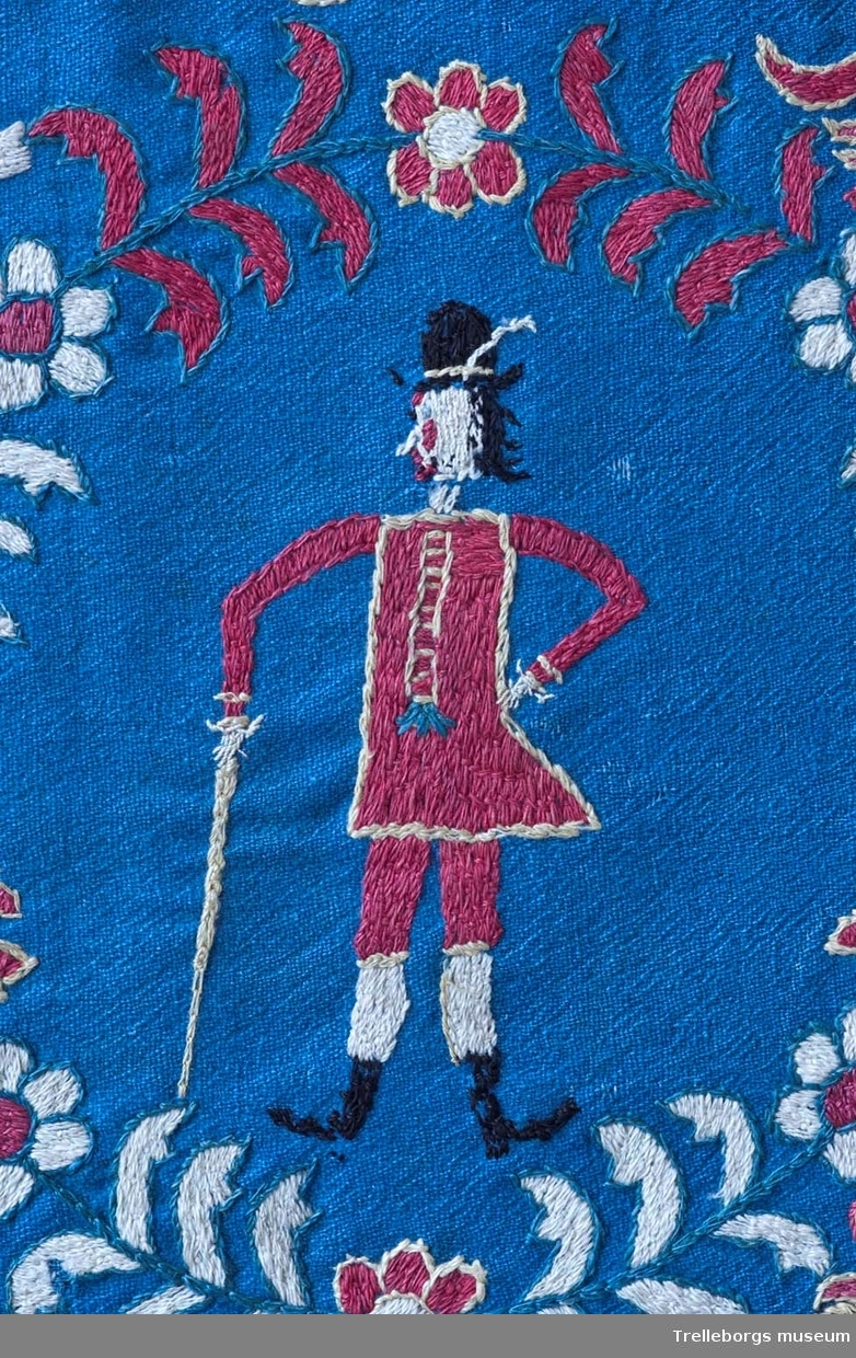 På en mellanblå botten är två kransar broderade, i den ena kransen står en kvinna och i den andra en man. I dynans hörn är blommor broderade, samma blomma kommer igen i dynans mitt tillsammans med en tupp, två gäss och två kor. Broderiets färger är rött, vitt, gult, grönt och svart. Dynan är märkt Anno 1780 Nils Hansson Ulgren Kierstin Andersdotter.