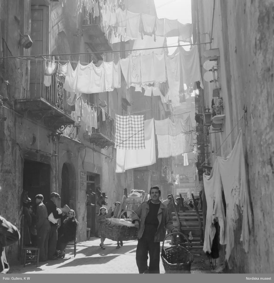 Gata i Neapel med folkvimmel och upphängd tvätt mellan husen. Utanför en port hänger skor till försäljning.