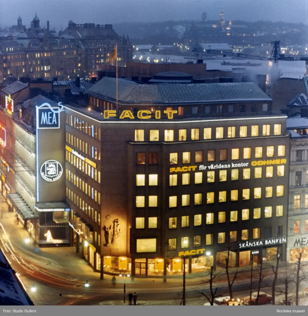 Stockholm. Kvällsvy över husen i hörnet Kungsträdgårdsgatan och Hamngatan, bland annat Åtvidabergshuset. På fasaden ljusskyltar för MEA, Facit och Skånska Banken. Vänstertrafik.