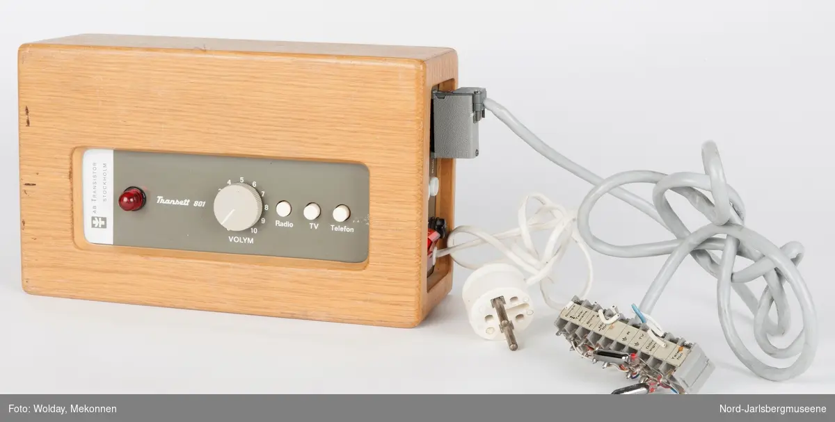 Firkantet apparat, som en liten radio. kassen er bygget av tre.
dsen kan kobles til telefon og radio, TV, eller brukes i forsamling med høreapparat. Antatt fra tidlig 1970-tall.
