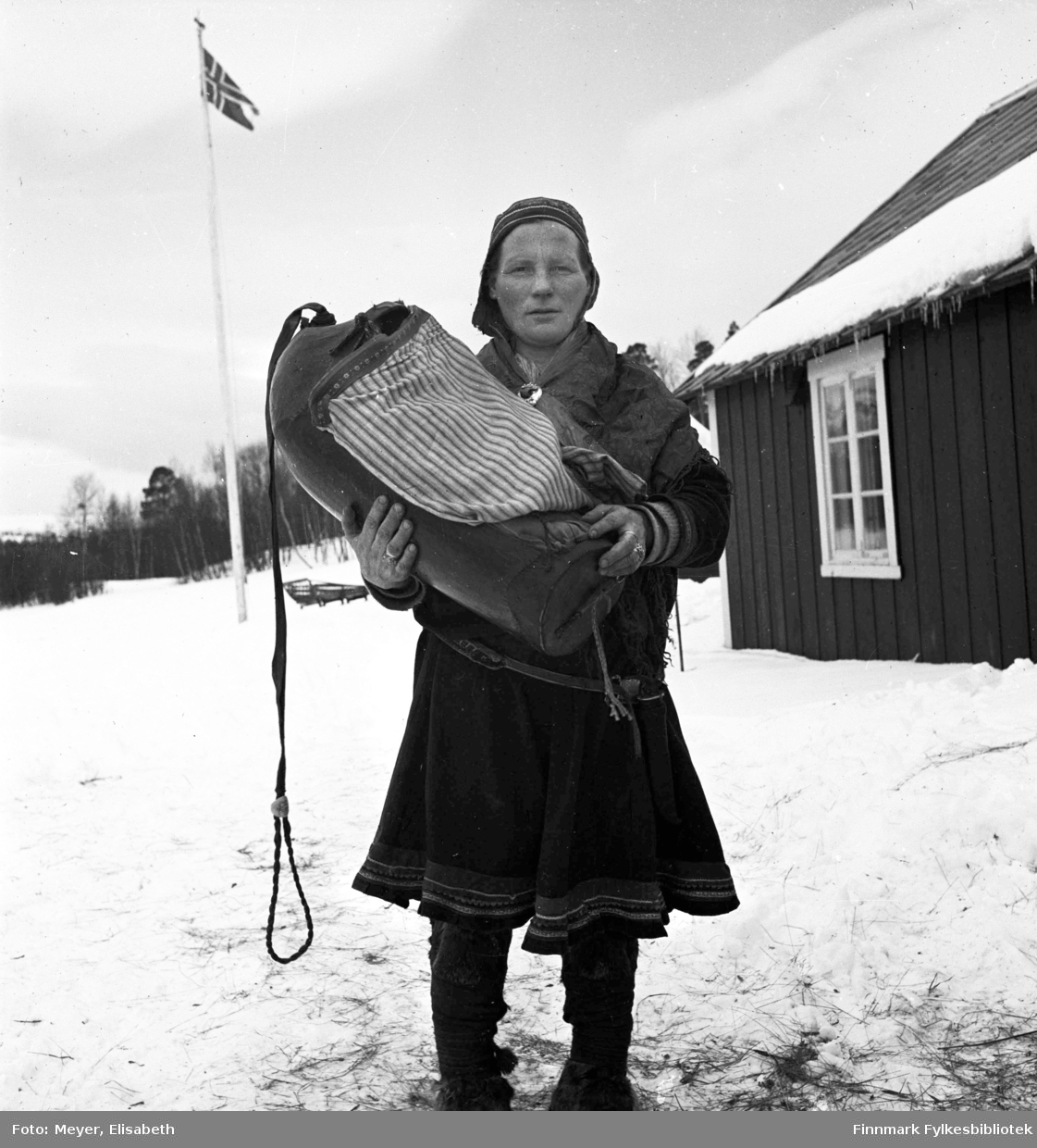 Susanne Aslaksdatter Siri med barnet i komse, utenfor en av Statens Fjellstuer. Muligens er dette Gargia Fjellstue. I bakgrunnen et norsk splittflagg på flaggstangen. Til høyre i bildet sees en bygning.