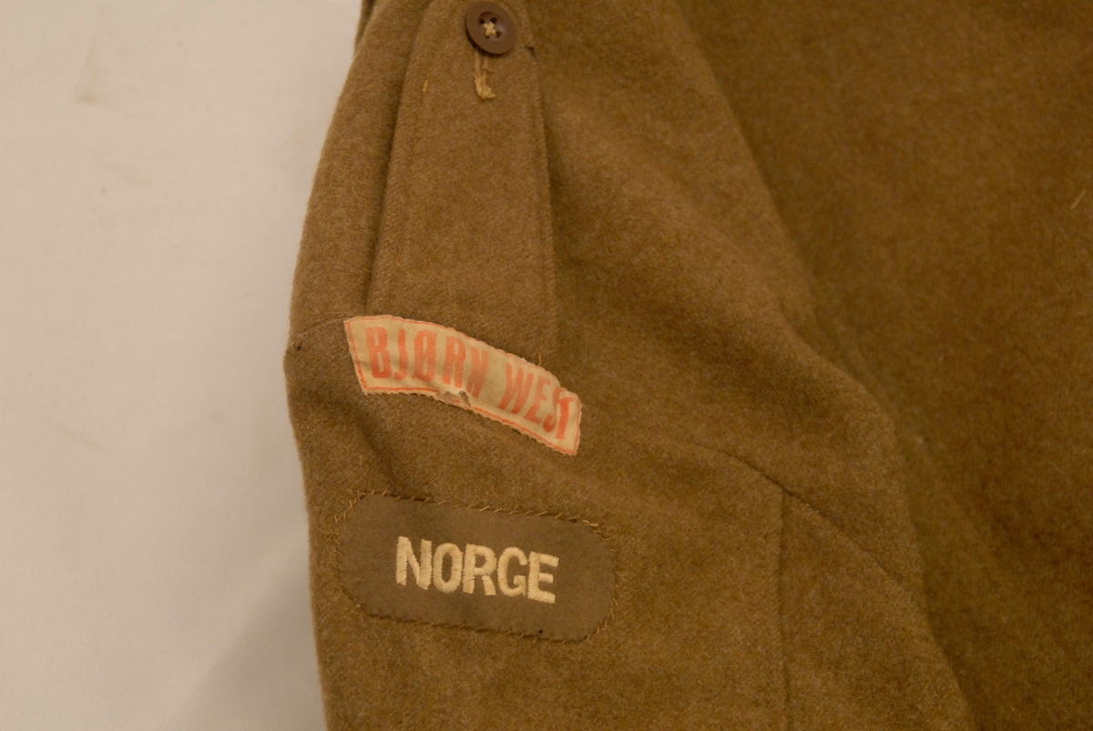 Lys brun battlejakke i ull med påsydde merker på armar. Brystlommer, knapper i front, på armar og lommer. Metallspenne nederst.