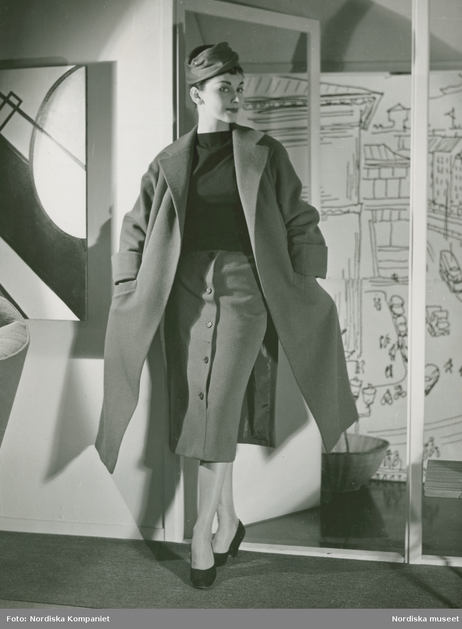 Modell i mörk tröja, kjol, kappa och hatt, framför en dörr. Konstverk på väggen. Modellbutiken.